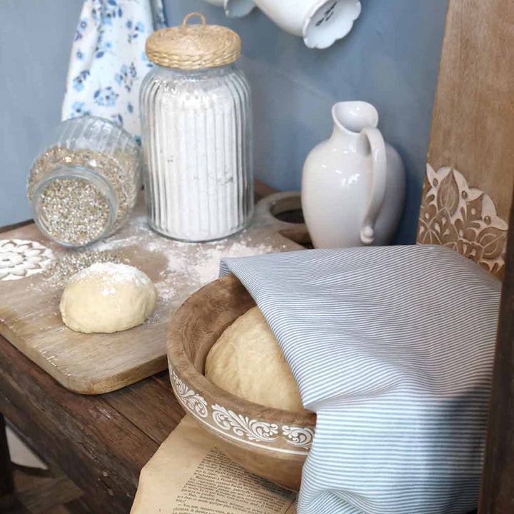 Eine Holzschüssel mit Teig, bedeckt mit dem Chic Antique - Geschirrtuch Maritimes Streifenmuster, steht auf einem rustikalen Tisch neben Teig, Mehl, Körnern in einem Glas, einem Krug und einer Tasse.