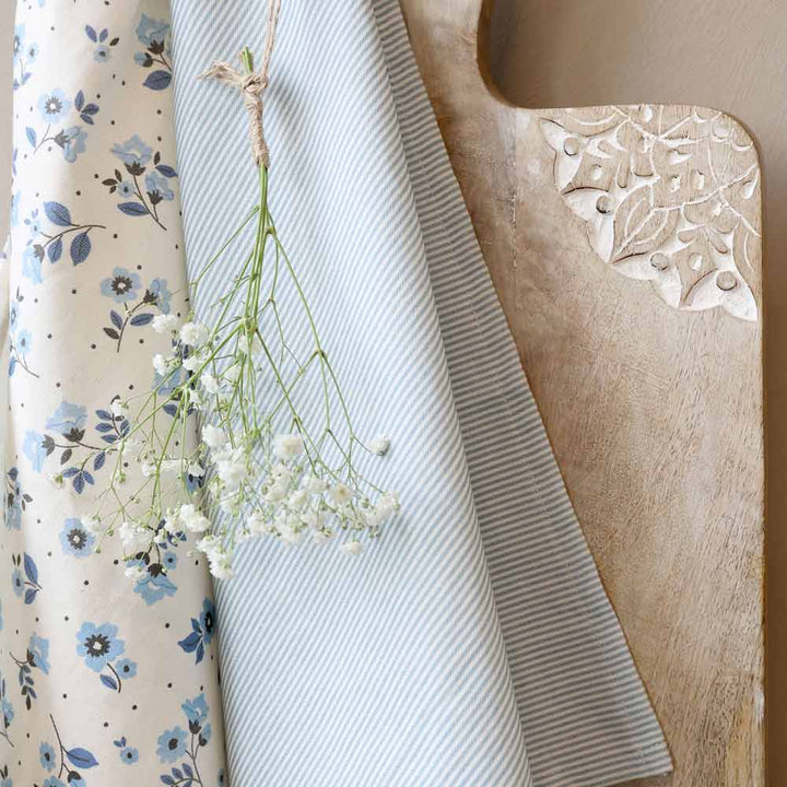 Das Chic Antique - Geschirrtuch Maritimes Streifenmuster in hellblauen Streifen wird neben einer geblümten Stoffrolle und einem Holzbrett mit geschnitzter Ecke präsentiert, über dem weiße Blumen hängen.