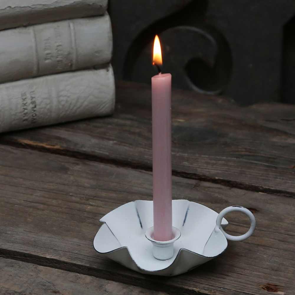 Eine brennende rosafarbene Kerze im Halter Chic Antique Kammerleuchter für Spitzkerzen White steht auf einer hölzernen Oberfläche, mit gestapelten Büchern im Hintergrund.