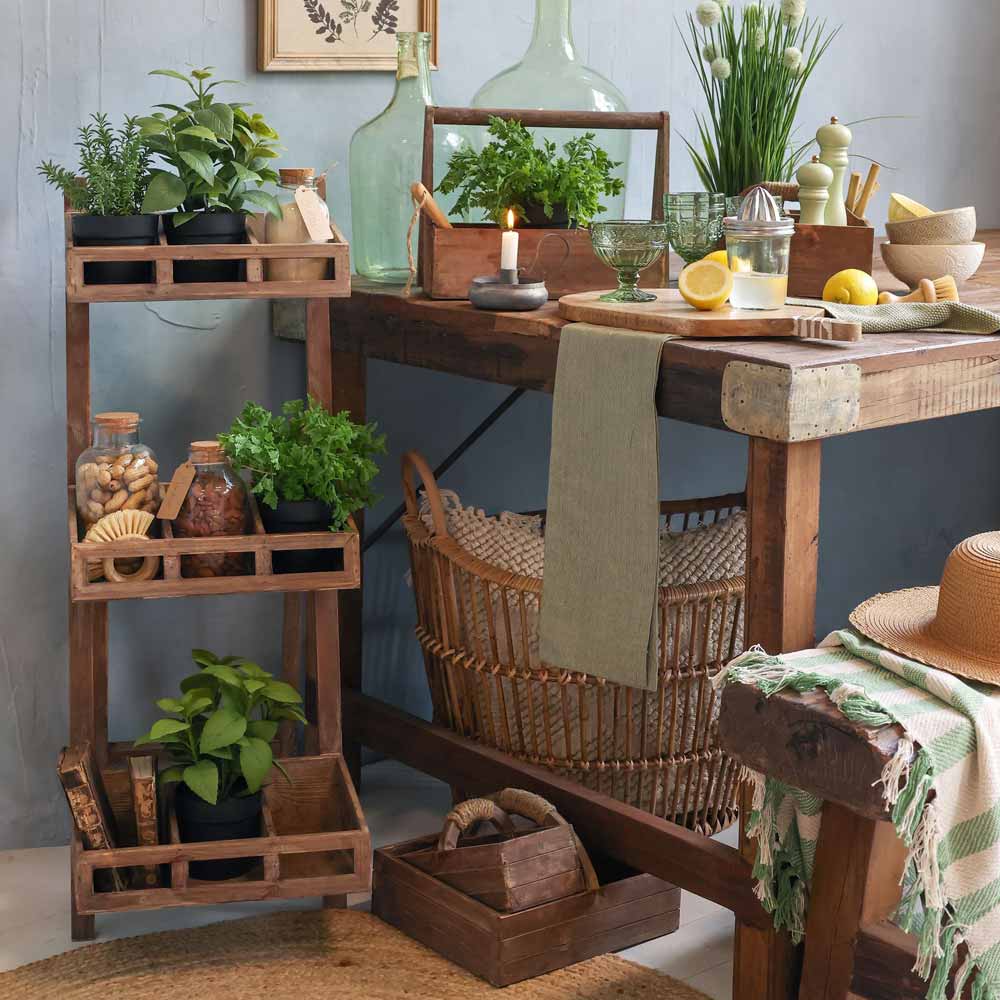 Eine rustikale Küchenszene zeigt die Holzregale von Chic Antique mit Kräutertöpfen und Gläsern. Der Tisch ist mit Glasflaschen, einer brennenden Kerze, Zitronen und dem geflochtenen Korb „Kasten mit Handgriff und 2 Kisten“ von Chic Antique geschmückt.