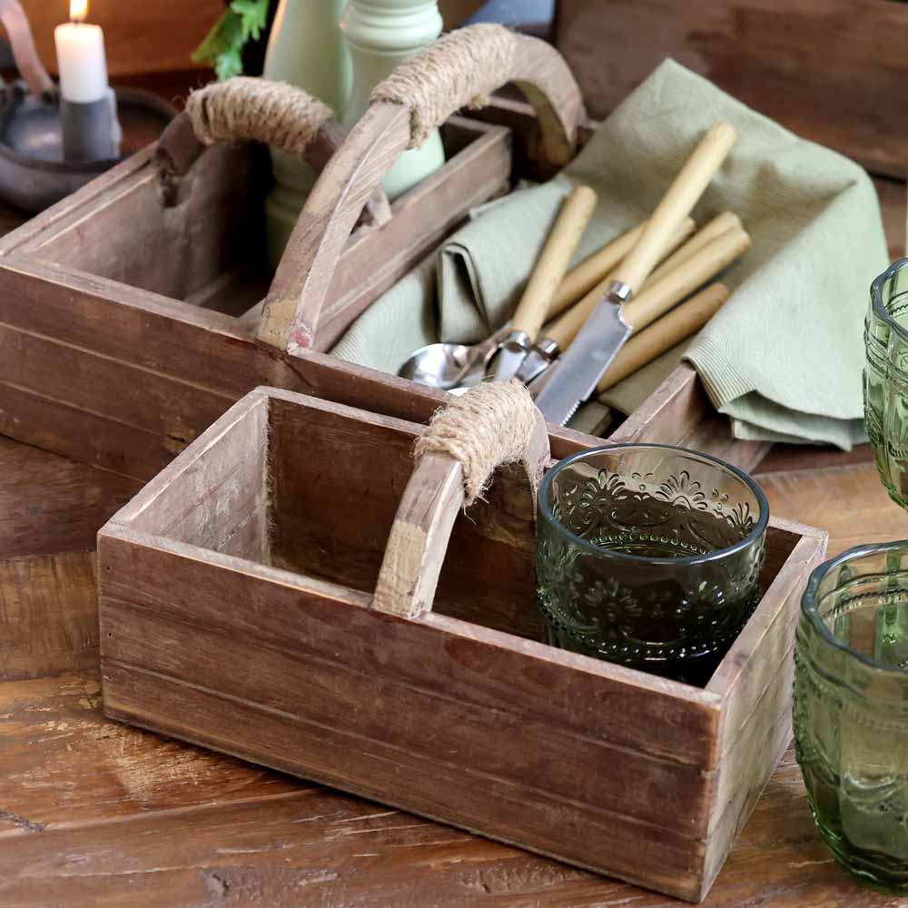 Auf einem Tisch stehen zwei Chic Antique – Kasten mit Handgriff und zwei Kisten aus Holz mit Seilgriffen. Eine Kiste enthält Besteck auf Stoff, die andere ein grünes Glas. Im Hintergrund befinden sich Kerzen und eine Holzoberfläche.