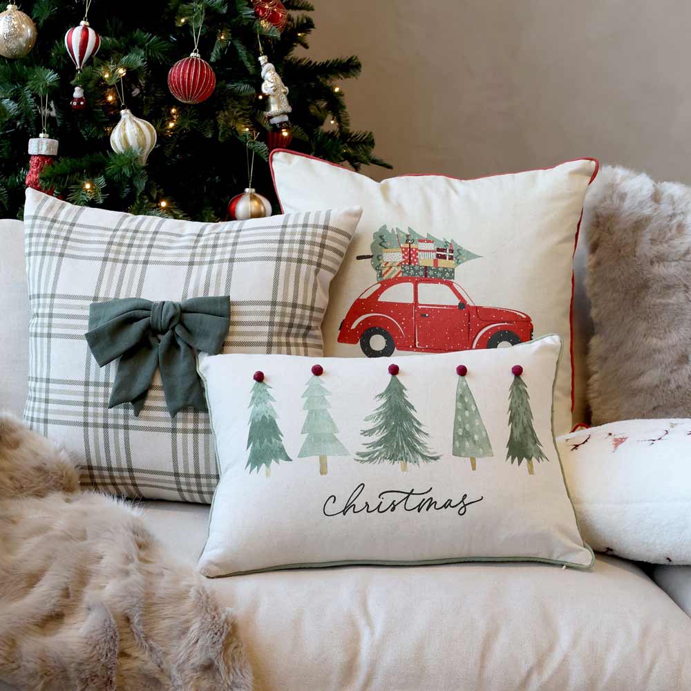 Auf einem Sofa liegen zwei dekorative Weihnachtskissen, eines davon ist der Chic Antique - Kissebezug Weihnachtsauto Leinen 50 x 50 cm, der ein rotes Auto mit einem Weihnachtsbaum zeigt. Ein weiteres Kissen hat eine Schleife und das dritte zeigt Bäume mit dem Wort „Weihnachten“. Im Hintergrund sorgt ein Weihnachtsbaum für festliche Stimmung.