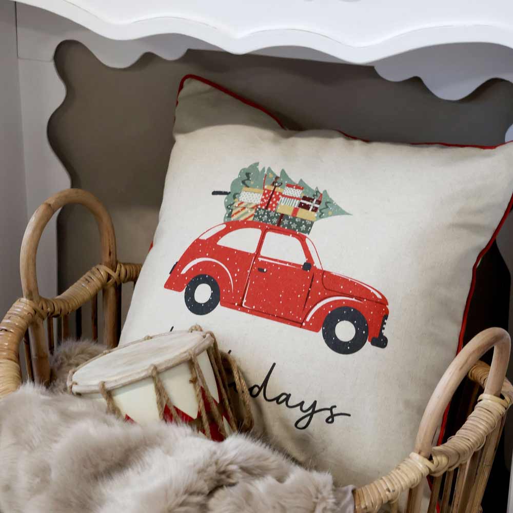 Ein Chic Antique - Kissebezug Weihnachtsauto Leinen 50 x 50 cm Kissen mit der Abbildung eines roten Autos, das einen Weihnachtsbaum und Geschenke transportiert, eingebettet in einen Weidenkorb neben einer Pelzdecke und einer dekorativen Trommel.