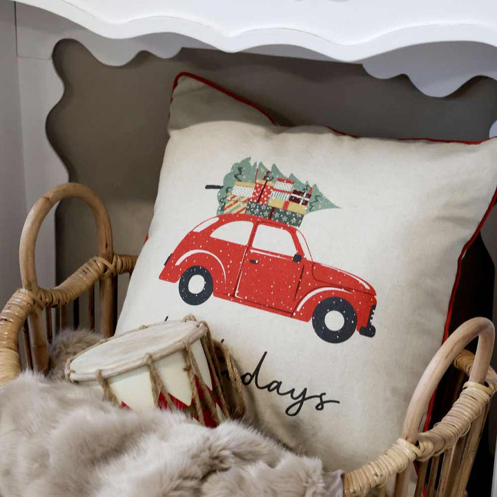 Ein Chic Antique - Kissebezug Weihnachtsauto Leinen 50 x 50 cm Kissen mit der Abbildung eines roten Autos, das einen Weihnachtsbaum und Geschenke transportiert, eingebettet in einen Weidenkorb neben einer Pelzdecke und einer dekorativen Trommel.