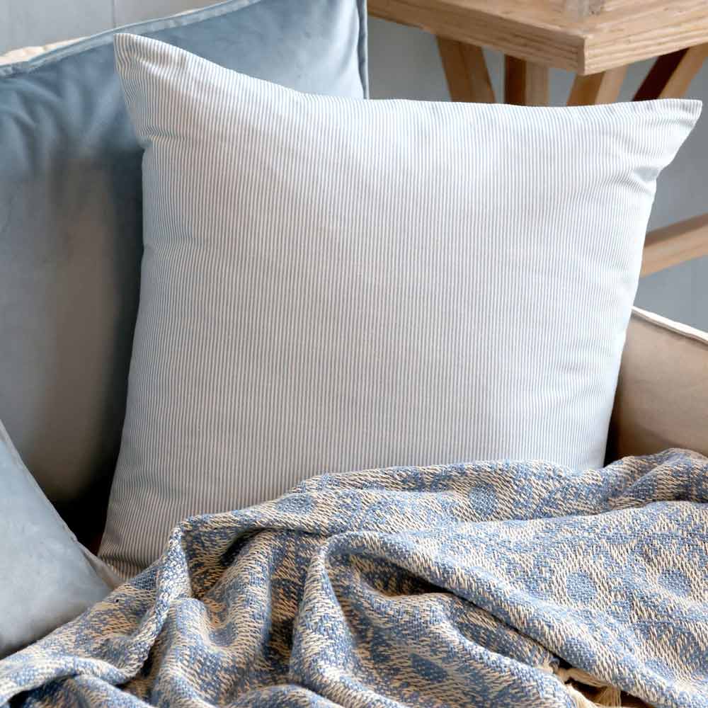 Der Chic Antique Kissenbezug Maritimes Streifenmuster in Hellblau liegt auf einem hellen Sofa neben einer blau gemusterten Decke, im Hintergrund steht ein Beistelltisch aus Holz.
