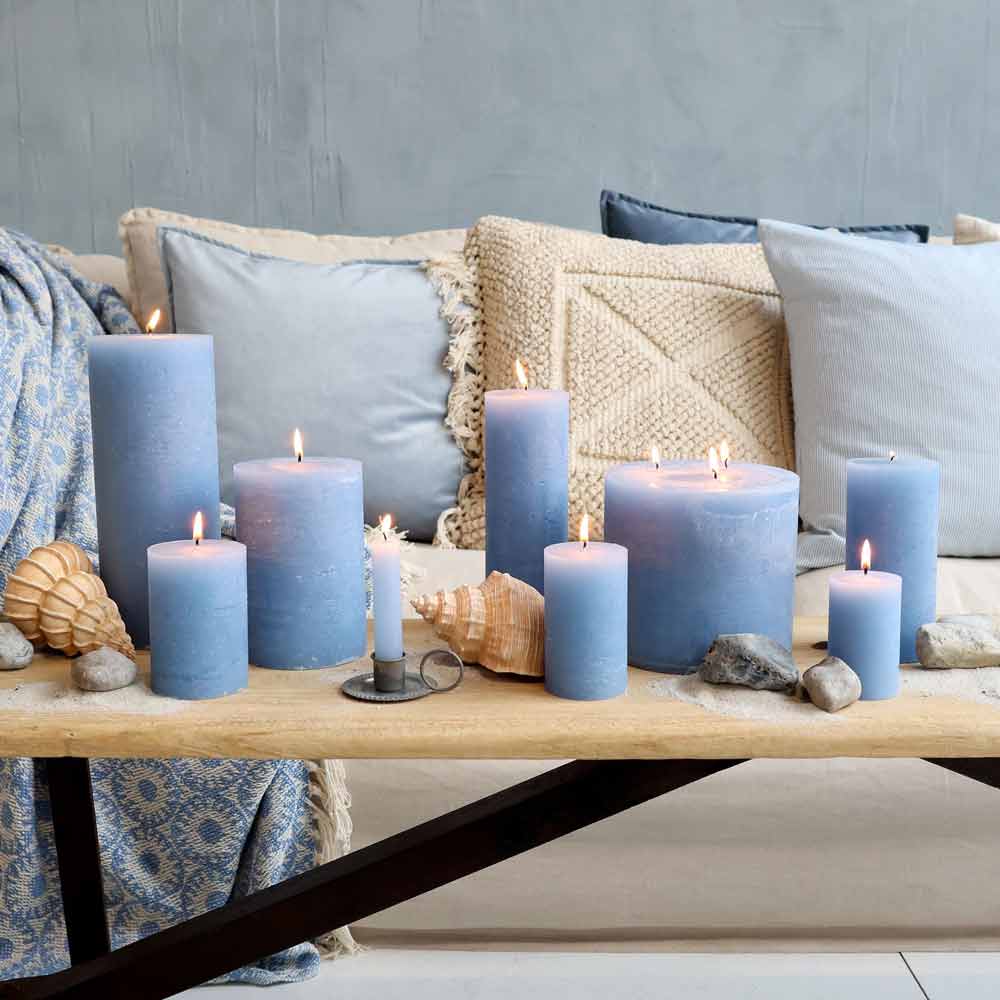 Auf einem Holztisch stehen brennende blaue Stumpenkerzen, Muscheln und Steine, im Hintergrund steht ein beiges Sofa, das mit dem Kissenbezug „Maritimes Streifenmuster“ von Chic Antique dekoriert ist.