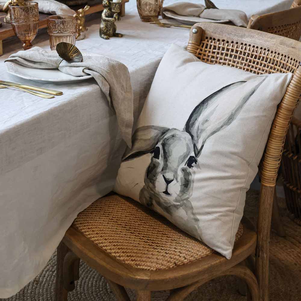 Ein Esstisch mit einer weißen Tischdecke ist mit Gläsern und Servietten gedeckt. Auf einem Stuhl liegt ein Kissen mit dem 45 x 45 cm großen Leinenbezug „Kissenbezug mit Hase Männchen“ von Chic Antique.