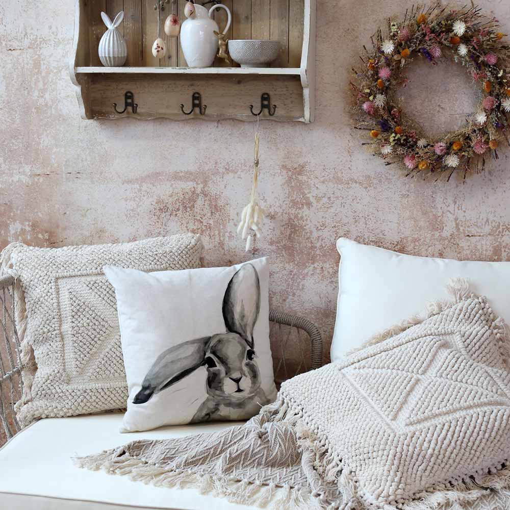 Eine gemütliche Sitzecke mit weißen und beigen Kissen, darunter der Chic Antique Kissenbezug mit Hase Männchen Leinen 45 x 45 cm. Darüber befindet sich ein Wandregal, geschmückt mit Dekoartikeln und einem Kranz aus Trockenblumen.