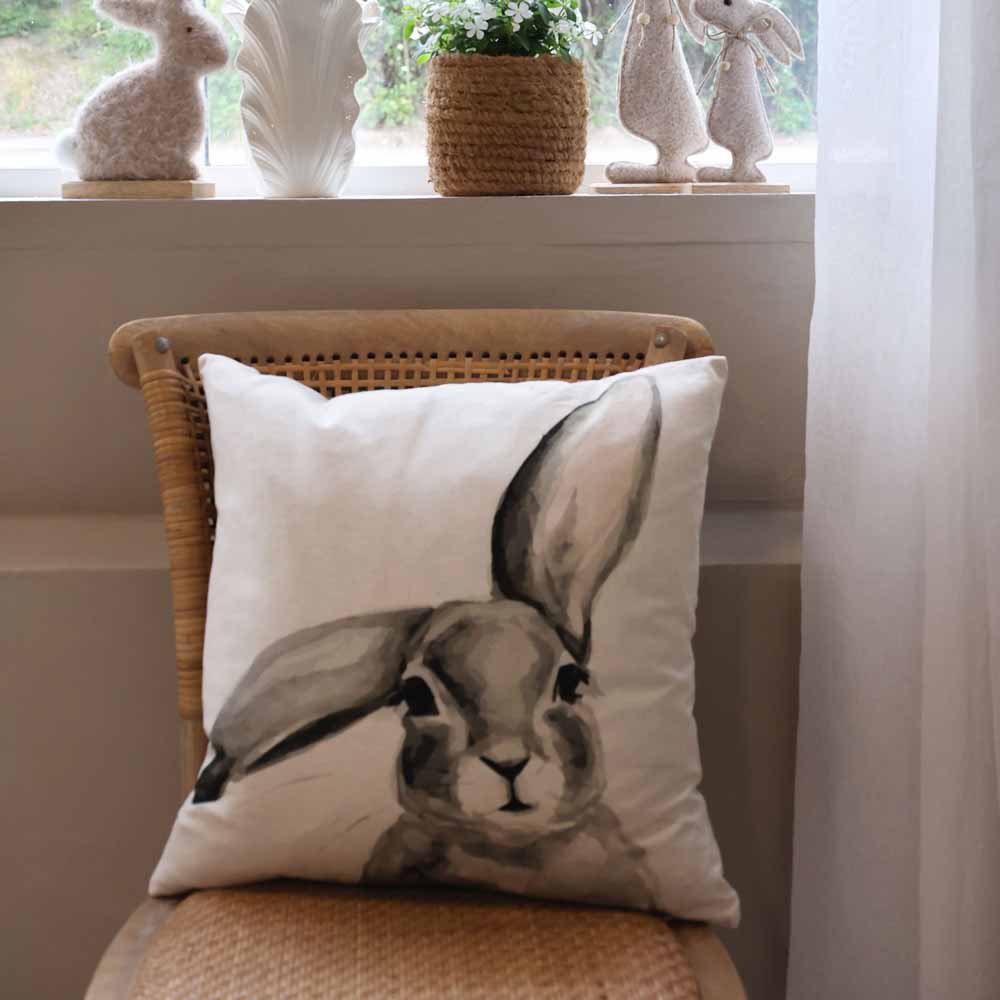 Ein Leinenkissenbezug von Chic Antique mit einer monochromen Hasenillustration liegt auf einem geflochtenen Stuhl. Dahinter schmücken dekorative Hasenfiguren und eine kleine Topfpflanze die Fensterbank.