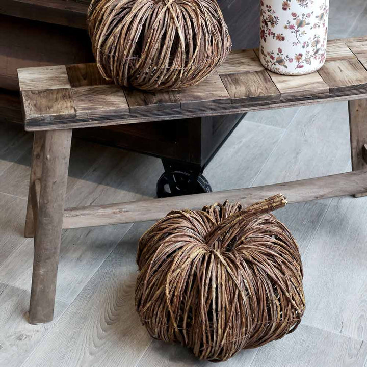 Zwei Chic Antique - Kürbis Rattan-Kürbisse aus geflochtenem Rattan schmücken eine rustikale Holzbank, die neben einer Vase mit Blumenmuster auf einem hellen Holzboden steht.