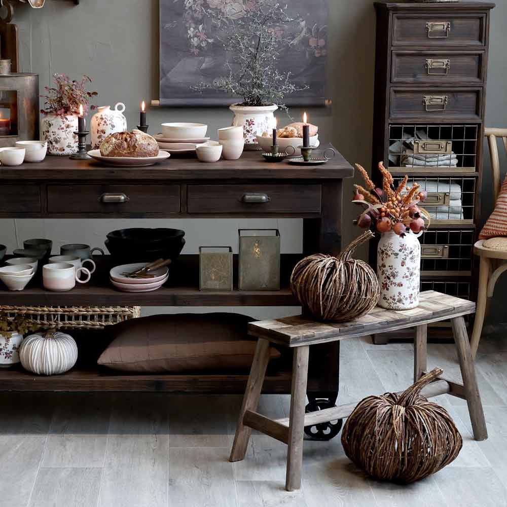 Eine rustikale Küchenszene mit dunklen Holzmöbeln, Keramikgeschirr, Kerzen, Trockenblumen und dekorativen geflochtenen Kürbissen von Chic Antique – Kürbis Rattan.