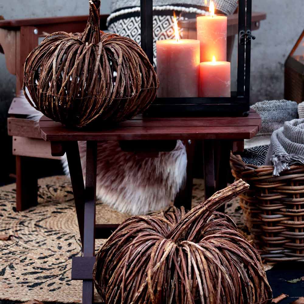 Zwei Chic Antique - Kürbis Rattan-Kürbisse stehen auf einer Holzbank neben einer Laterne mit brennenden Kerzen, einem geflochtenen Korb und Textilien.