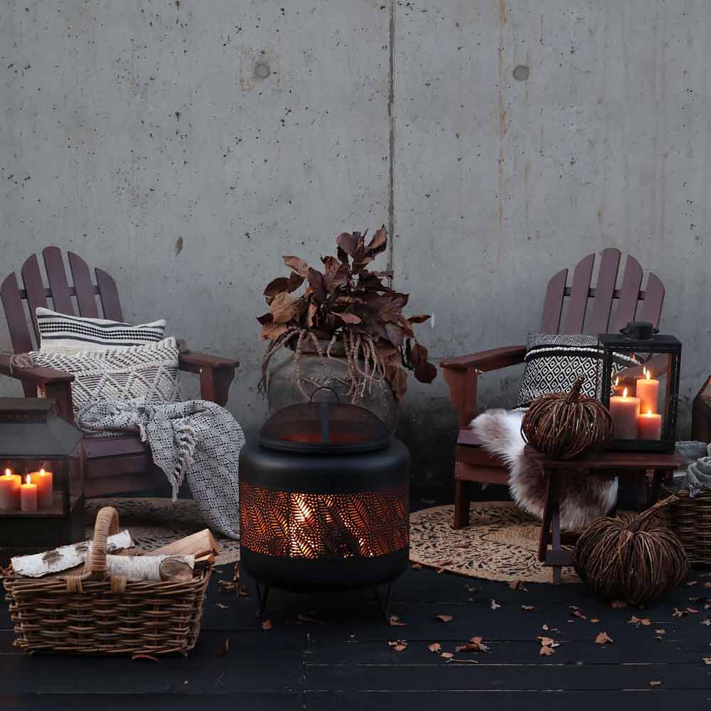 Zwei Chic Antique Kürbis-Rattanstühle mit Kissen und Decken stehen neben einer kleinen Feuerstelle, umgeben von Kerzen und einem Korb mit Brennholz vor einer Betonwand.