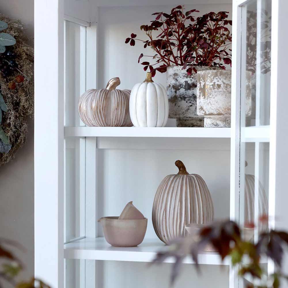 Auf weißen Regalen stehen Chic Antique Kürbis mit Gold Antique Creme, ein kleiner Krug mit Deckel und Topfpflanzen mit roten Blättern.