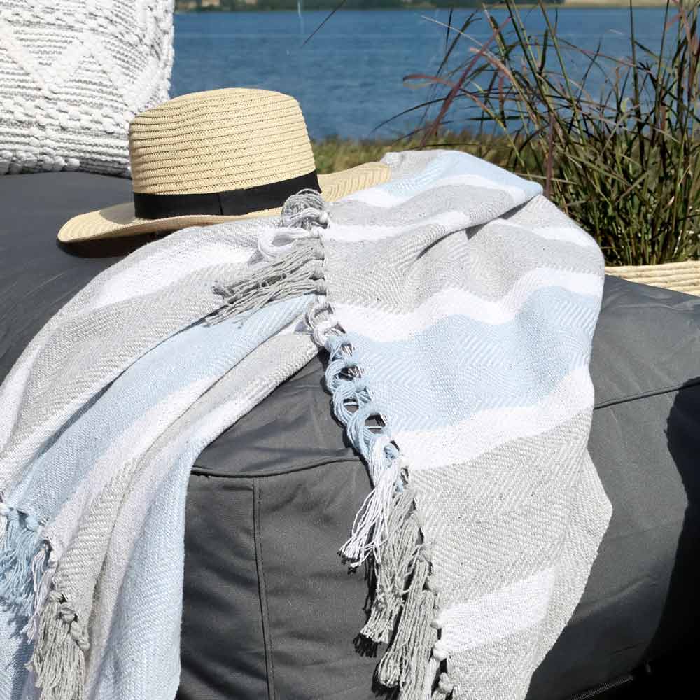 Die karierte Kuscheldecke „Grau-Blau mit Fransen“ von Chic Antique ist neben einem Strohhut auf einem grauen Outdoor-Kissen drapiert, mit Blick auf den See und das Gras im Hintergrund.