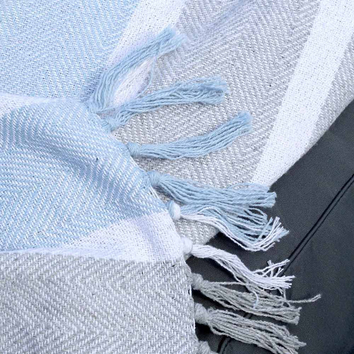 Eine Nahaufnahme der Chic Antique – Plaid Kuscheldecke Grau-Blau mit Fransen, mit einem hellblauen und grauen Fischgrätenmuster mit Fransen, das teilweise über eine dunkelgraue Oberfläche drapiert ist.