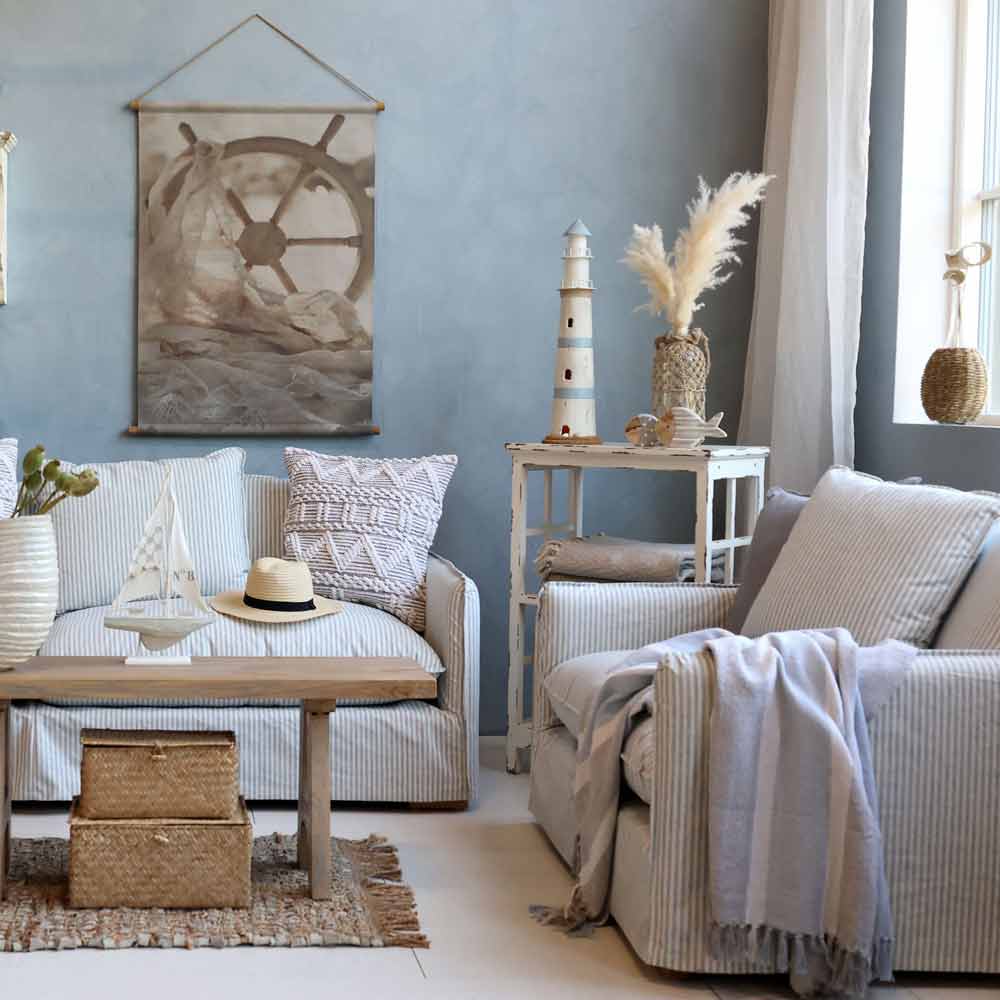 Wohnzimmer im Küstenstil mit der Plaid-Kuscheldecke Grau-Blau mit Fransen von Chic Antique, einem gestreiften Sofa und Sessel, nautischem Dekor wie einem Leuchtturmmodell, Wandkunst mit Schiffssteuerrad, Weidenkörben und neutralen Akzenten.