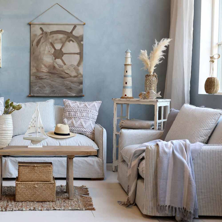 Wohnzimmer im Küstenstil mit der Plaid-Kuscheldecke Grau-Blau mit Fransen von Chic Antique, einem gestreiften Sofa und Sessel, nautischem Dekor wie einem Leuchtturmmodell, Wandkunst mit Schiffssteuerrad, Weidenkörben und neutralen Akzenten.