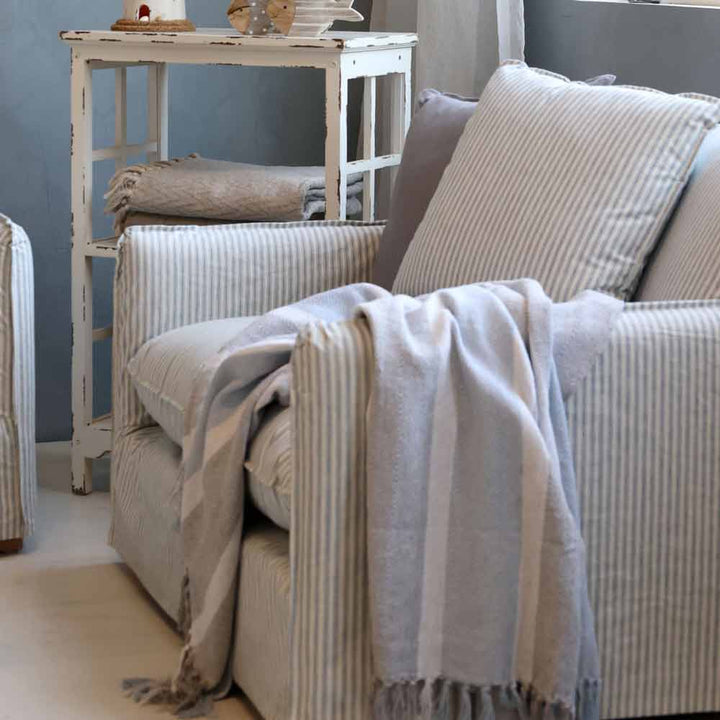 Ein gestreifter Sessel mit einer darüber drapierten Chic Antique – Plaid Kuscheldecke in Grau-Blau mit Fransen steht neben einem kleinen weißen Beistelltisch aus Holz, auf dem gefaltete Decken und Dekoartikel liegen.