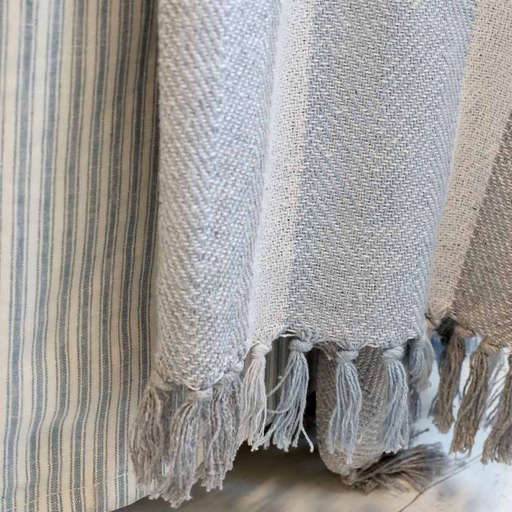 Nahaufnahme der Chic Antique – Plaid Kuscheldecke Grau-Blau mit Fransen, die einen weichen graublauen Webstoff mit Fransenenden und einer Mischung aus Chevron- und Streifenmustern zeigt.