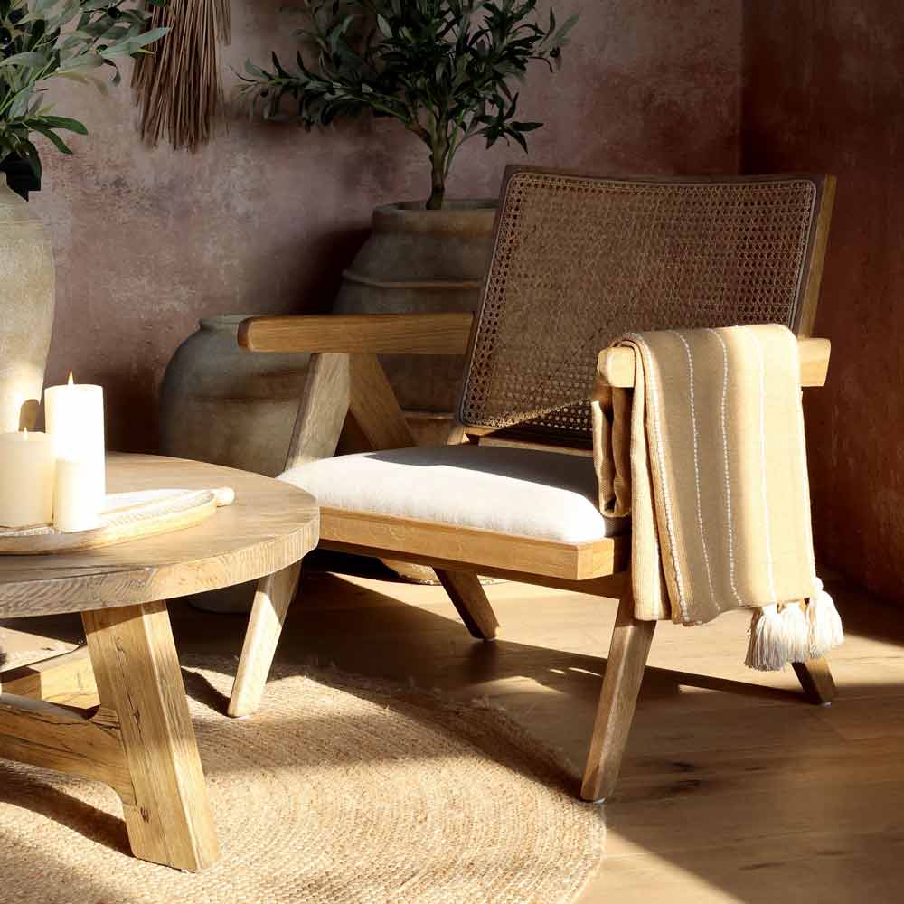 Neben einem runden Holztisch mit Kerzen steht ein Loungesessel aus Holz mit geflochtener Rückenlehne und weißem Kissen; über dem Sessel ist das Chic Antique - Plaid mit Fransen im Naturbraun-Boho-Look drapiert.