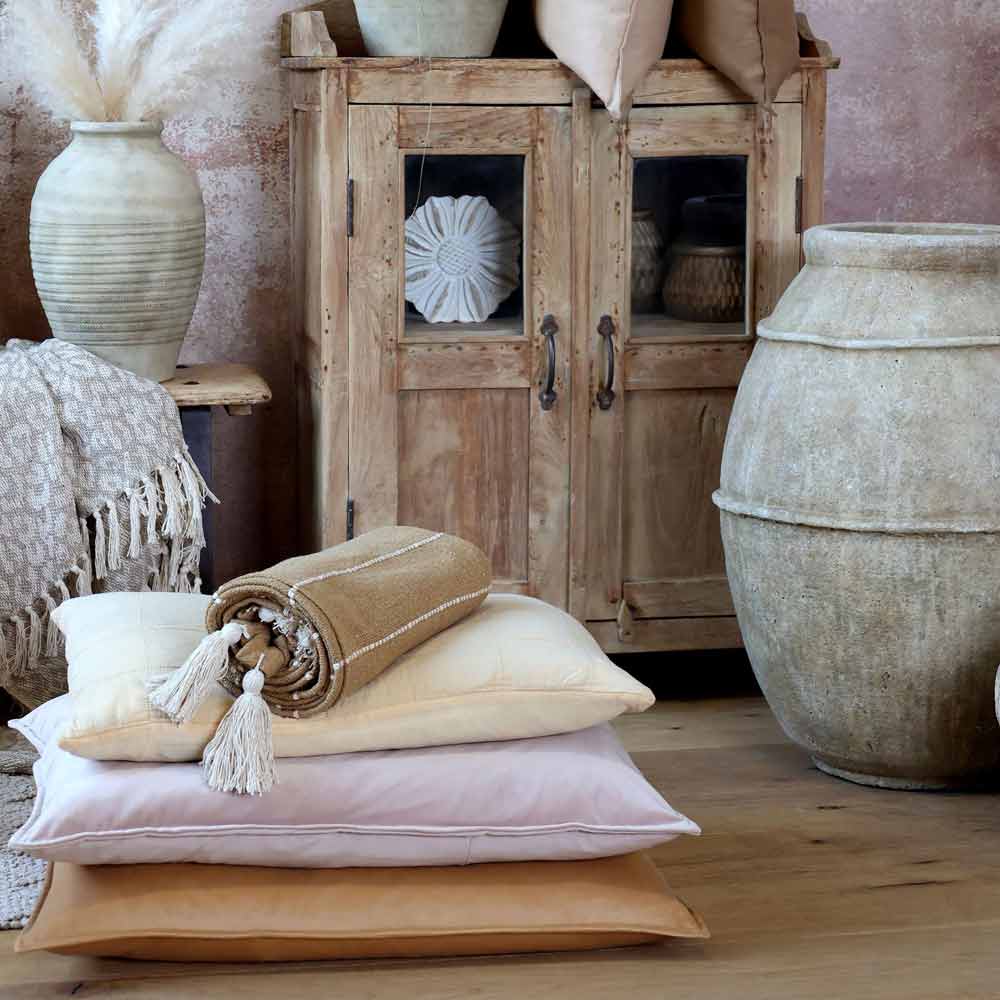 Ein Stapel Kissen mit dem darauf gefalteten Chic Antique - Plaid mit Fransen in Naturbraun Boho Look liegt auf einem Holzboden neben einer großen Keramikvase, einem Schrank und dekorativer Keramik in einem rustikalen Raum.