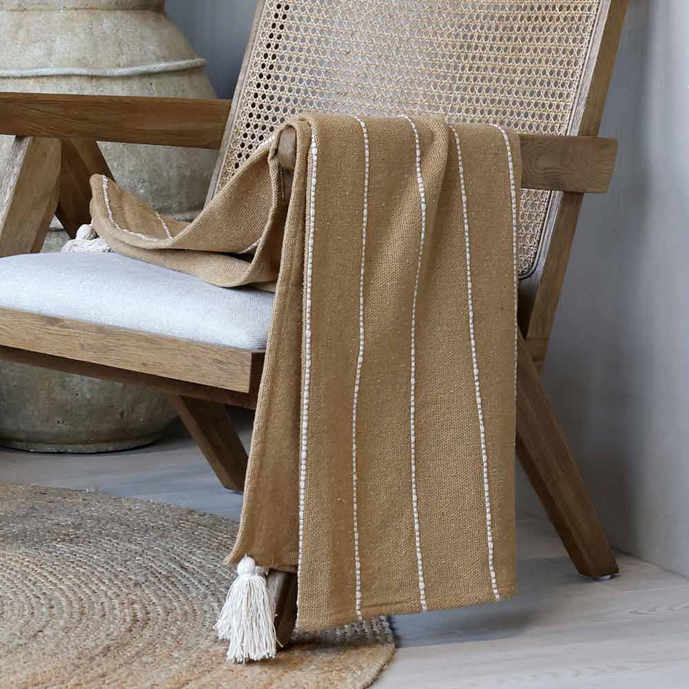 Auf einem Holzstuhl mit geflochtener Rückenlehne steht das Chic Antique Plaid mit Fransen im Naturbraun-Boho-Look, daneben ein runder gewebter Teppich und eine große Keramikvase.