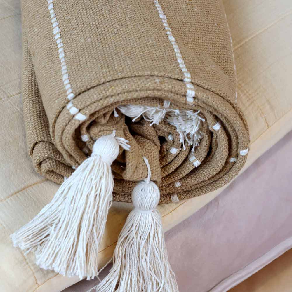 Das Chic Antique – Plaid mit Fransen in Naturbraun Boho Look, eine beige gewebte Decke mit weißen Quasten und dünnen weißen Streifen, liegt auf einer hellen Oberfläche.