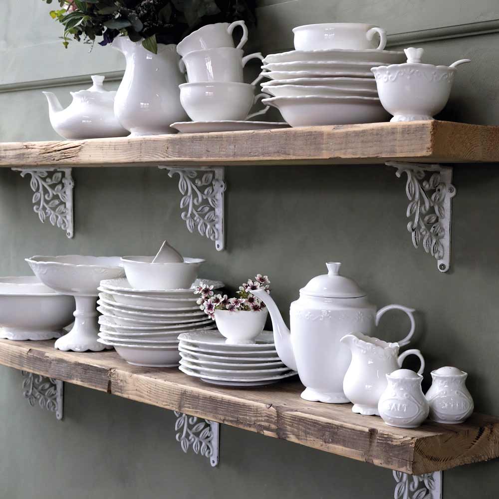 Auf Holzregalen ist weißes Porzellangeschirr ausgestellt, darunter Teller, Schüsseln, Tassen, eine Chic Antique - Provence Kanne 1 Liter, ein Milchkännchen und Zuckerdosen, alles ordentlich angeordnet. Dekorative Metallhalterungen stützen die Regale.
