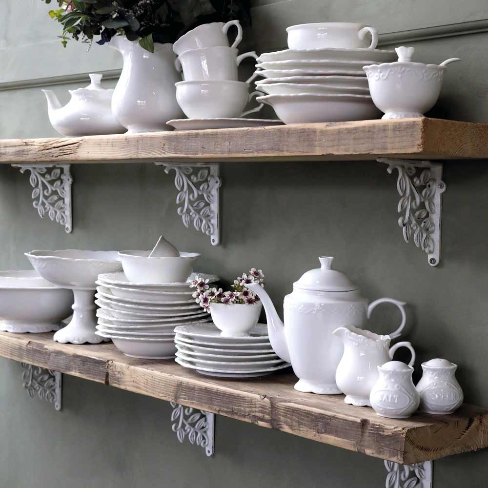 Auf zwei an der Wand montierten Holzregalen sind verschiedene weiße Keramikgeschirre, darunter Teller, Schüsseln, Krüge und ein Chic Antique - Provence Milchkännchen, ordentlich angeordnet. Zwischen einigen Geschirrteilen stehen kleine dekorative Pflanzen.