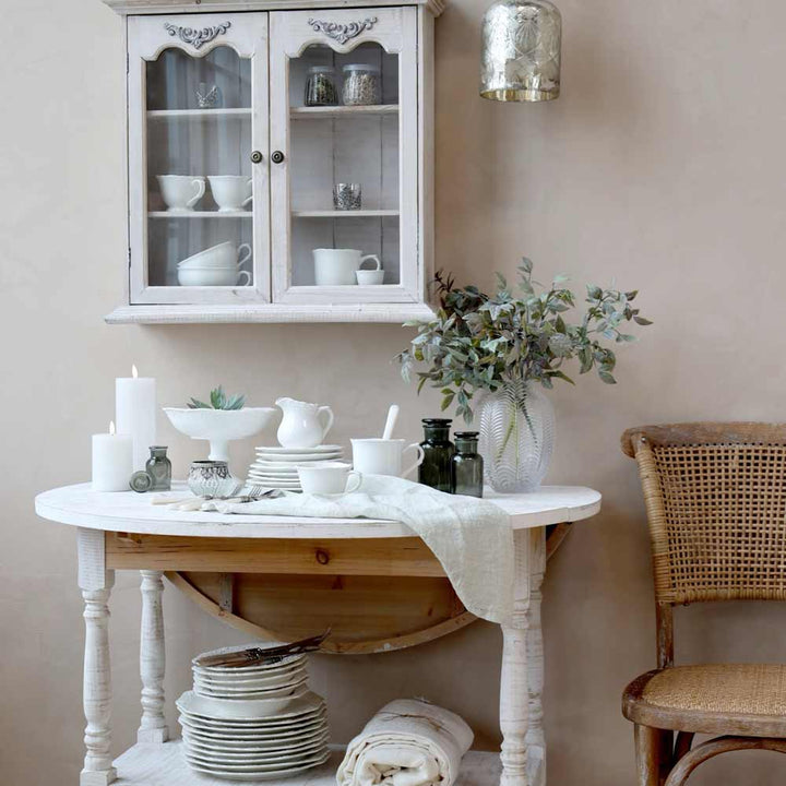 Eine rustikale Küchenecke mit einem weißen Tisch mit gestapeltem Geschirr, Kerzen, grünen Flaschen und einer Topfpflanze. Über dem Tisch befindet sich ein weißer Schrank mit Glastüren, auf dem Chic Antique - Provence Milchkännchen ausgestellt ist.