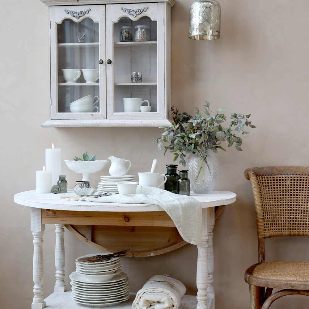 Ein weißer Konsolentisch aus Holz mit gestapelten Chic Antique - Provence Schüsseln mit Fuß, Kerzen und einer Pflanze steht unter einem Wandschrank mit weißem Geschirr. Neben dem Tisch steht ein Rattanstuhl.