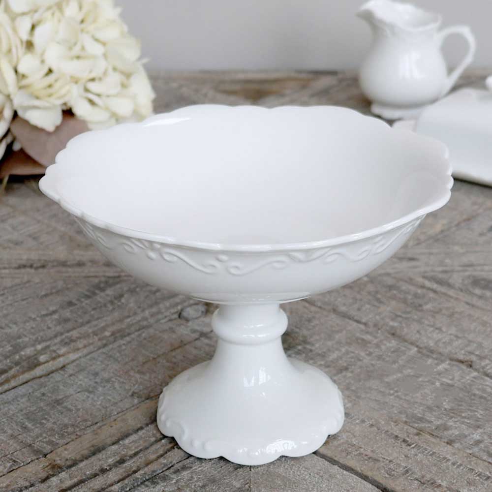 Eine Chic Antique - Provence Schüssel mit Fuß mit gewellten Rändern und dekorativen Details auf einer Holzoberfläche, begleitet von einem weißen Krug und weißen Hortensienblüten im Hintergrund.