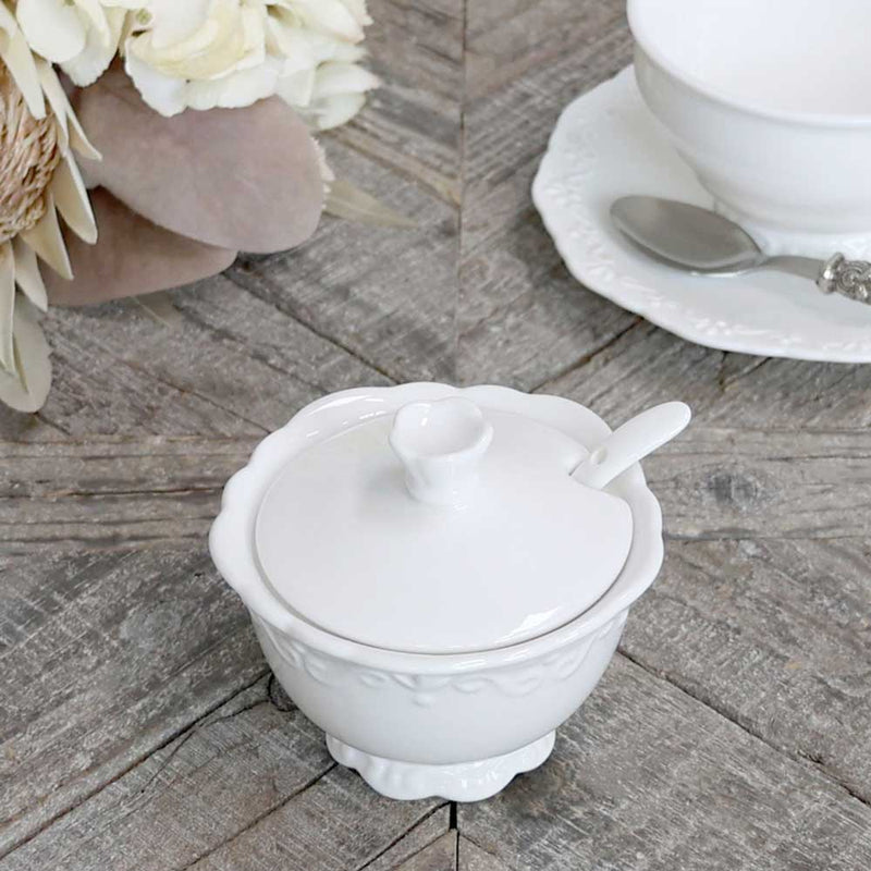 Eine Chic Antique - Provence Zuckerdose mit Löffel steht auf einer Holzoberfläche neben einer Untertasse, einem Löffel und Blumendekoration.