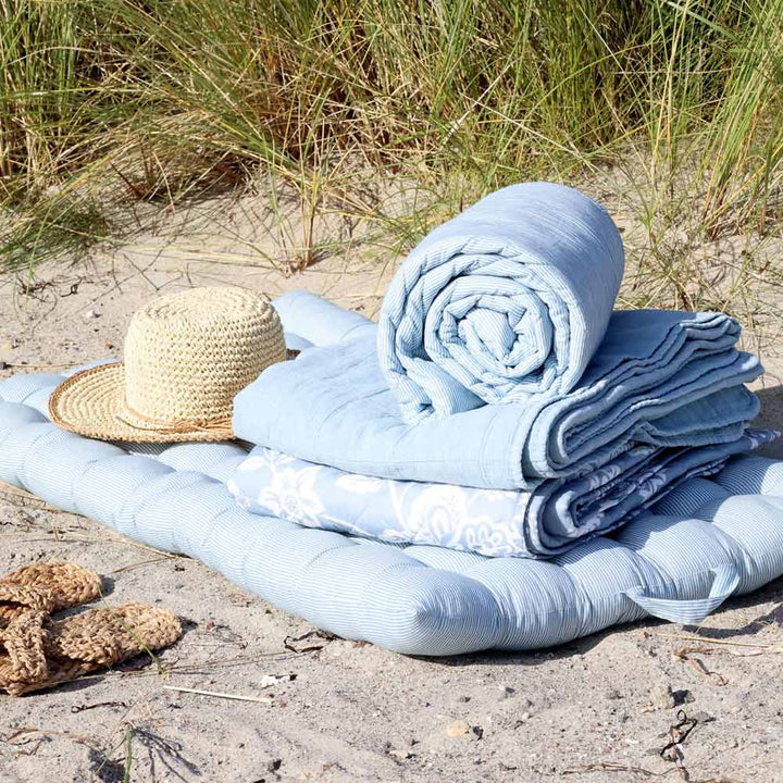 Der Chic Antique - Quilt Maritimes Streifenmuster Himmelblau 180 x 130 cm ist auf einer gepolsterten Matte auf sandigem Boden ausgebreitet, mit einer zusammengerollten Decke, einem Strohhut daneben und hohem Gras im Hintergrund.