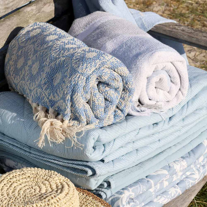 Der Chic Antique Quilt Maritimes Streifenmuster Sky Blue 180 x 130 cm wird mit einem weiteren hellblauen Quilt auf gefaltetem blauem Leinen präsentiert, während in der unteren linken Ecke ein gewebter Strohhut hervorschaut.