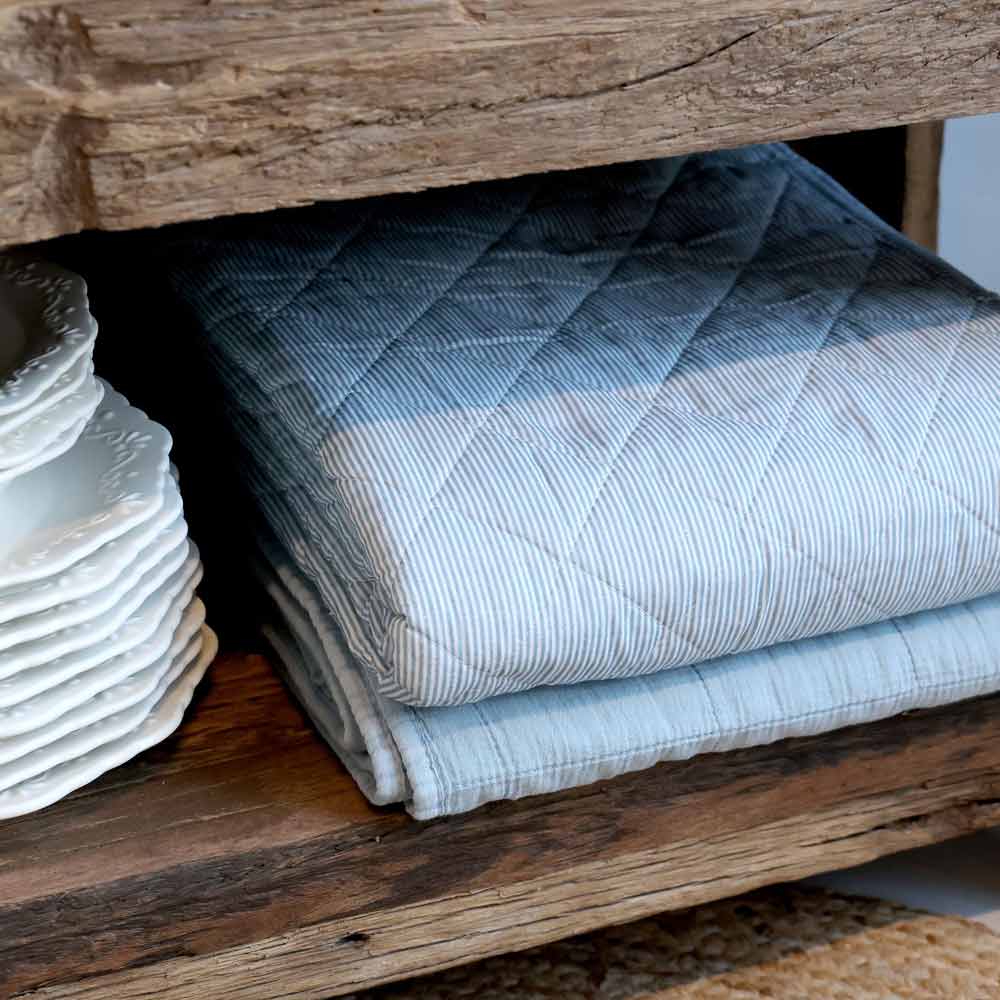 Der Chic Antique - Quilt Maritimes Streifenmuster Sky Blue 180 x 130 cm liegt gefaltet neben gestapelten weißen Keramiktellern auf einem Holzregal.
