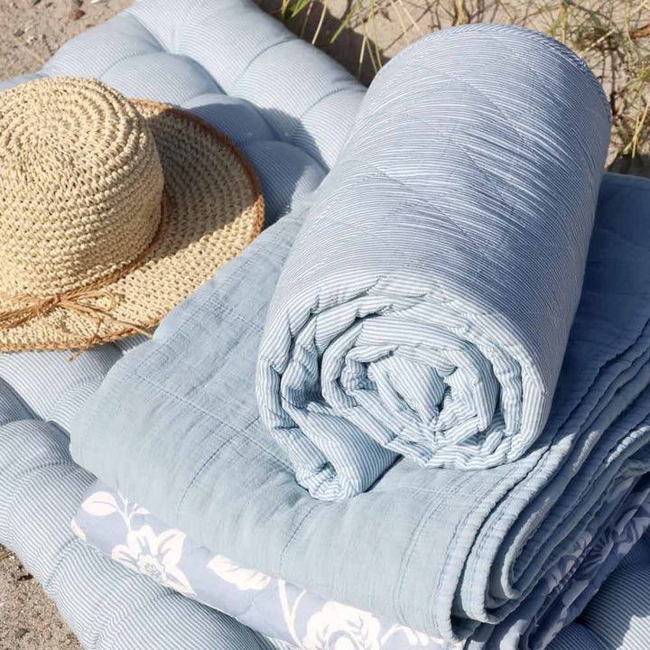 Ein Chic Antique Quilt Maritimes Streifenmuster Himmelblau 180 x 130 cm ist neben einer gerollten blau gestreiften Decke und einem Strohhut gefaltet, alles liegt im Freien auf einer gepolsterten Matte.