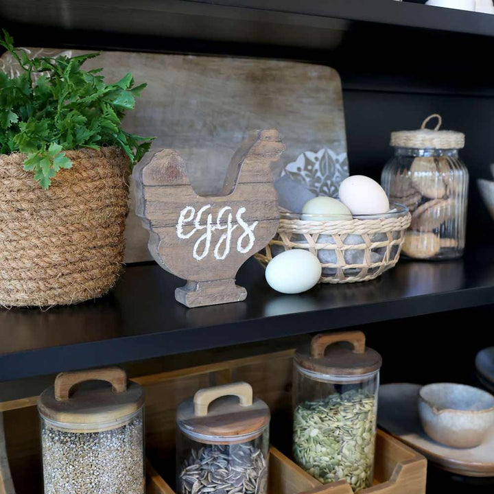 Auf einem Küchenregal sind neben verschiedenen Küchenutensilien eine Schüssel mit Flechtkorb von Chic Antique, eine Topfpflanze, ein hölzernes „Eier“-Schild, Glasbehälter mit Deckel und ein Korb mit Eiern ausgestellt.