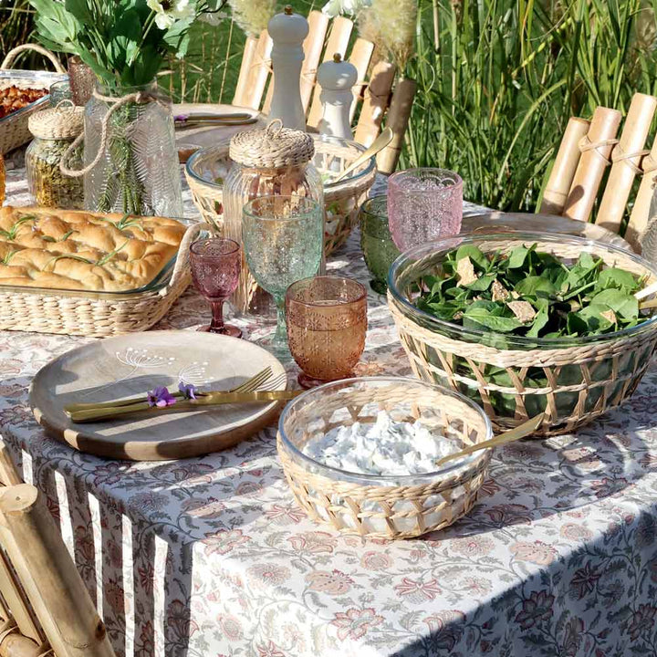 Gedeckter Tisch für draußen mit verschiedenen Gläsern, Tellern, Brot, Salat und der Chic Antique - Schüssel mit Flechtkorb auf einer geblümten Tischdecke mit Grünpflanzen im Hintergrund.