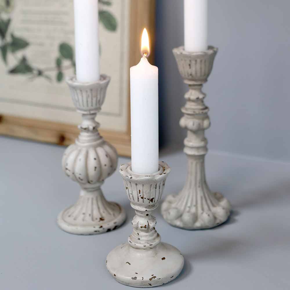 Drei Chic Antique – Shabby Chic Kerzenständer Antikgrau. Vintage-Kerzenständer mit brennenden weißen Kerzen sind auf einer hellen Oberfläche angeordnet, mit gerahmten Kunstwerken im Hintergrund.