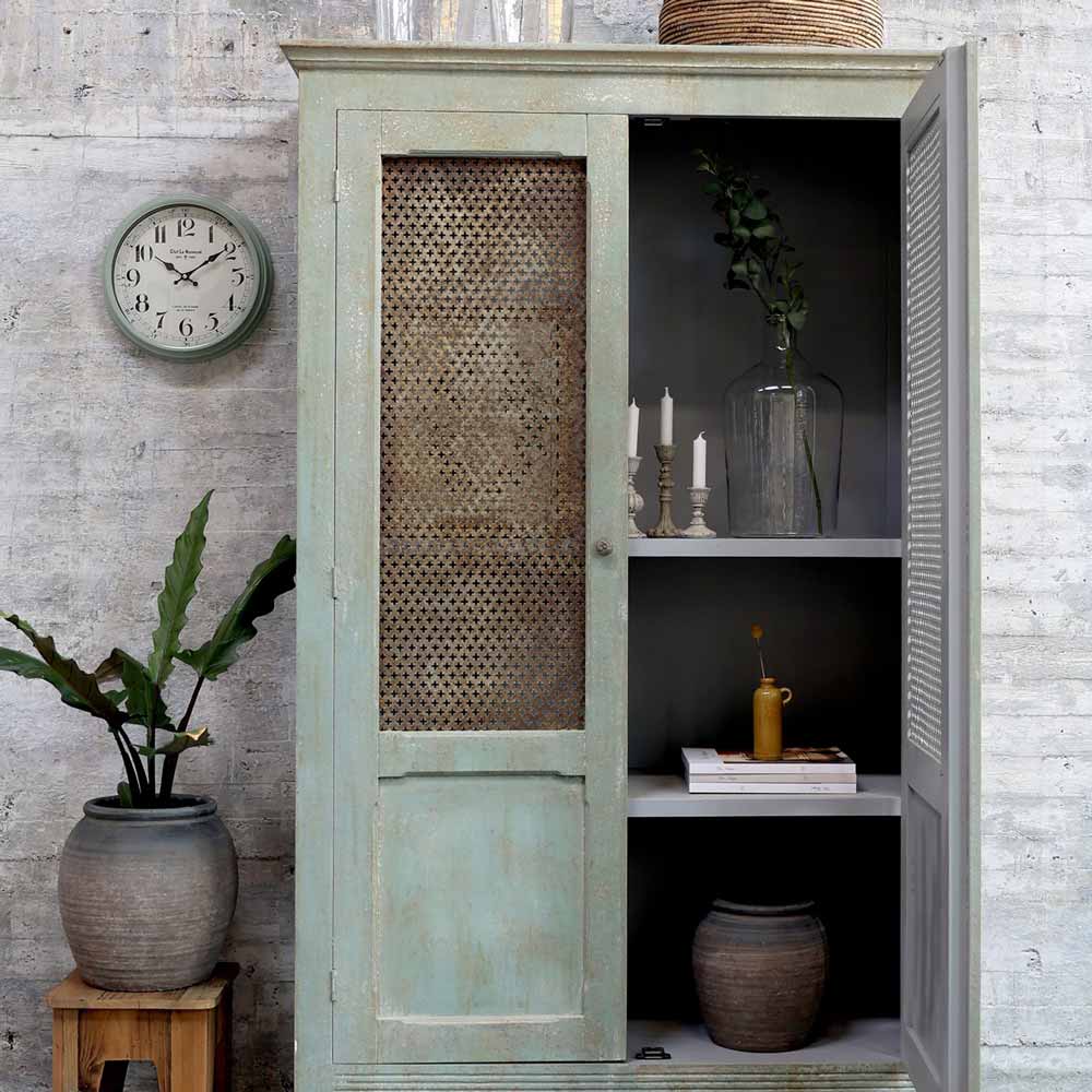 Ein hellgrüner Schrank im Used-Look mit offener Netztür stellt den Chic Antique - Shabby Chic Kerzenständer Antikgrau Vintage zwischen Kerzen, Büchern, Vasen und Pflanzen zur Schau; eine Wanduhr und Blumentöpfe stehen in der Nähe.