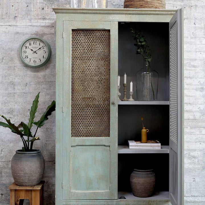 Ein hellgrüner Schrank im Used-Look mit offener Netztür stellt den Chic Antique - Shabby Chic Kerzenständer Antikgrau Vintage zwischen Kerzen, Büchern, Vasen und Pflanzen zur Schau; eine Wanduhr und Blumentöpfe stehen in der Nähe.