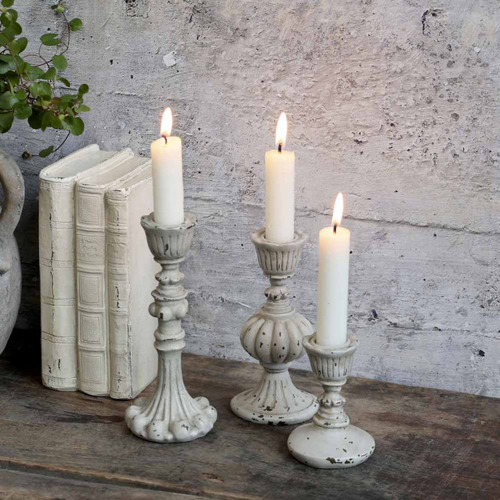 Drei brennende weiße Kerzen in den Kerzenständern „Chic Antique – Shabby Chic“ in Antikgrau (Vintage) stehen auf einer Holzoberfläche neben gestapelten weißen Büchern und einer kleinen Pflanze vor einer strukturierten grauen Wand.