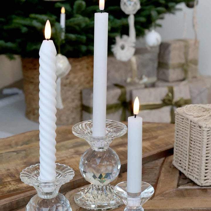 Drei Chic Antique - Stabkerze LED Twist 2er-Set unterschiedlicher Höhe in Glashaltern stehen auf einem Holztisch, im Hintergrund sind ein geschmückter Weihnachtsbaum und verpackte Geschenke zu sehen.
