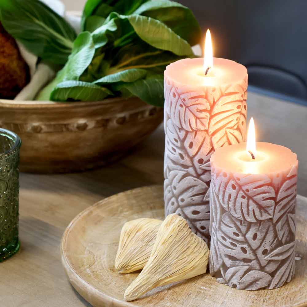 Zwei **Chic Antique – Stumpenkerze mit Blattmuster Macon** mit Blattmuster auf einem Holztablett, neben getrockneten Dekostücken und einer Schale mit Grünzeug im Hintergrund.