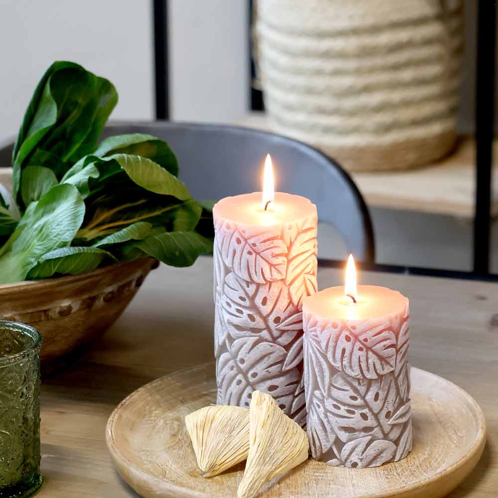 Zwei Chic Antique - Stumpenkerze mit Blattmuster Macon Kerzen auf einem Holztablett mit dekorativen Elementen, auf einem Tisch platziert. Daneben steht eine Schüssel Salat. Im Hintergrund ist ein geflochtener Korb zu sehen.