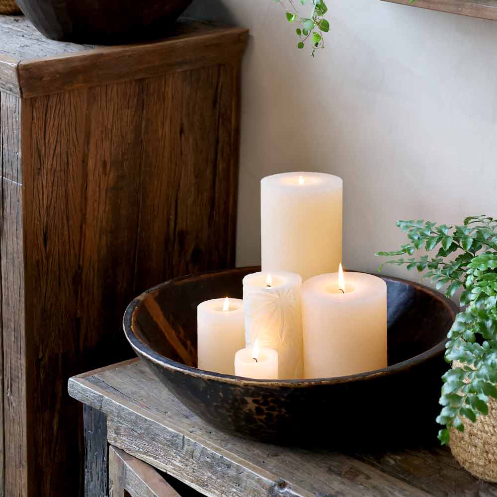 Eine Holzschale fasst fünf Chic Antique - Stumpenkerze mit Blattmuster Macon in verschiedenen Höhen, platziert auf einem rustikalen Holztisch neben einem Holzschrank und grünen Pflanzen.