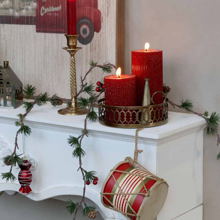 Ein weißer Kaminsims, dekoriert mit der gerippten und glitzernden Stumpenkerze von Chic Antique, einem kleinen Hausornament aus Metall, einer rot-weißen Trommel und einer Girlande mit roten und silbernen Kugeln.