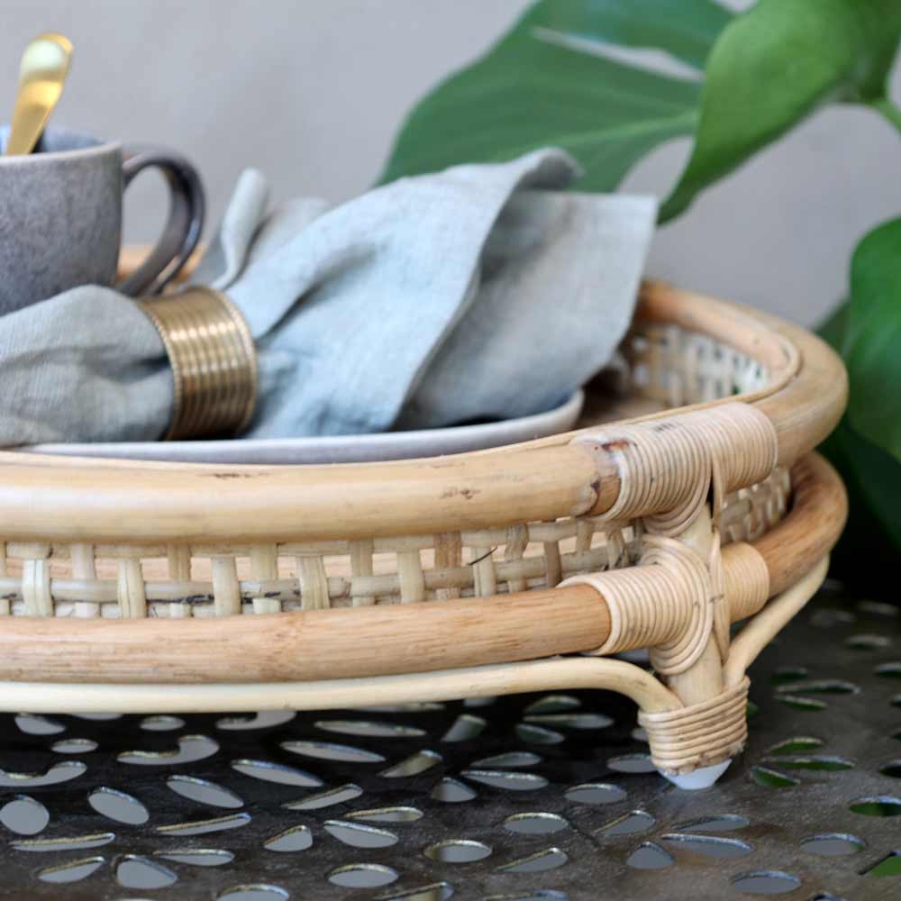 Ein Chic Antique - Tablett französisch geflochten auf Fuß mit einer grauen Tasse und einer Stoffserviette, mit grünen Blättern im Hintergrund.